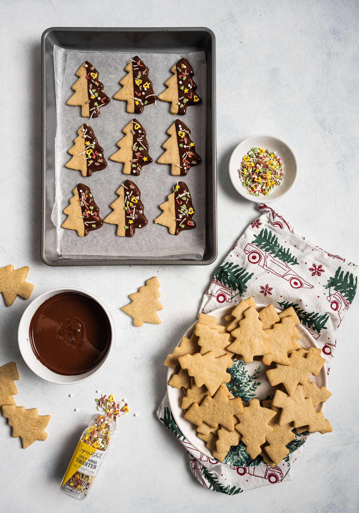 tree shaped cinnamon shortbread cookies half dipped in chocolate with sprinkles on baking sheet  plate of tree shaped cookies bowl of sprinkles bowl of melted chocolate