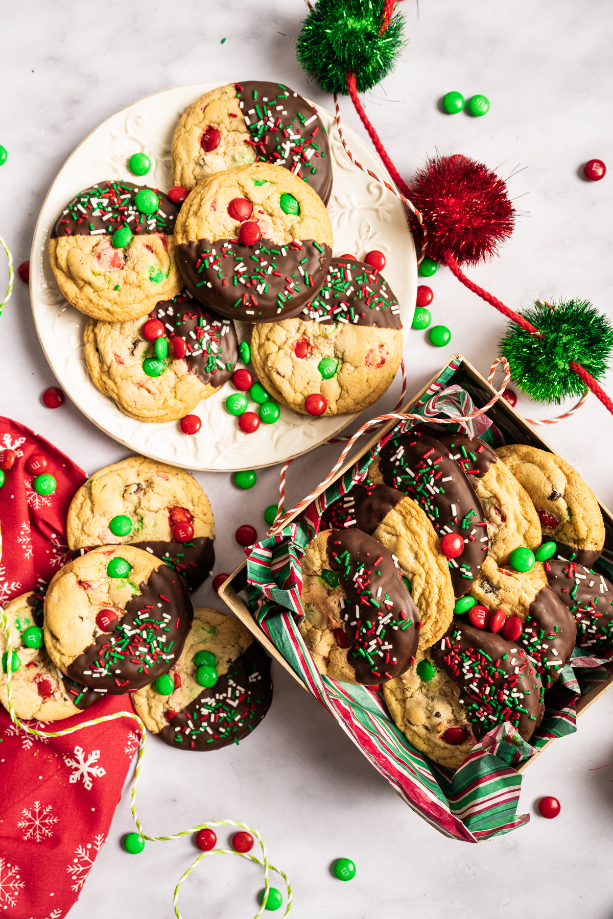 red and green M&M chewy chocolate chip cookies half dipped in chocolate decorated with red white and green sprinkles in box and on plate