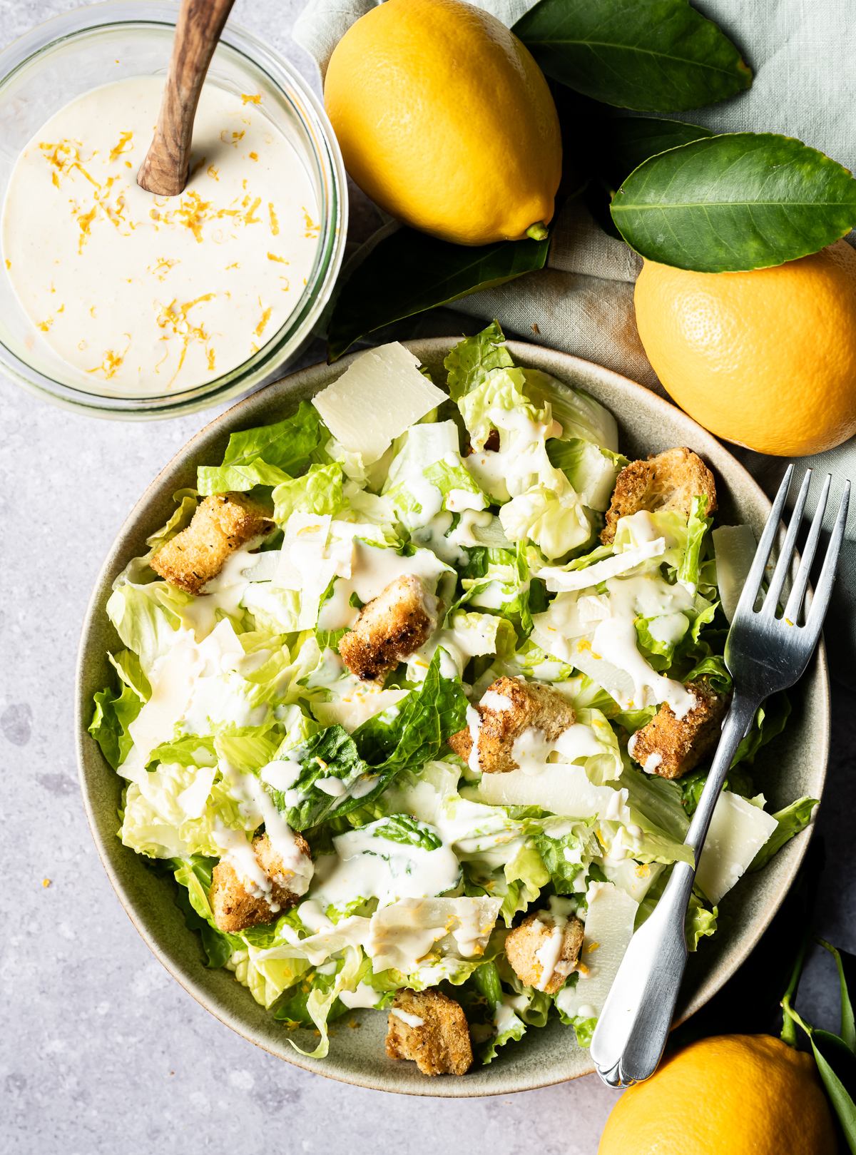 Bowl of lettuce with croutons shaved Parmesan lemon zest and Caesar dressing drizzled over it two whole meyer lemons jar of dressing