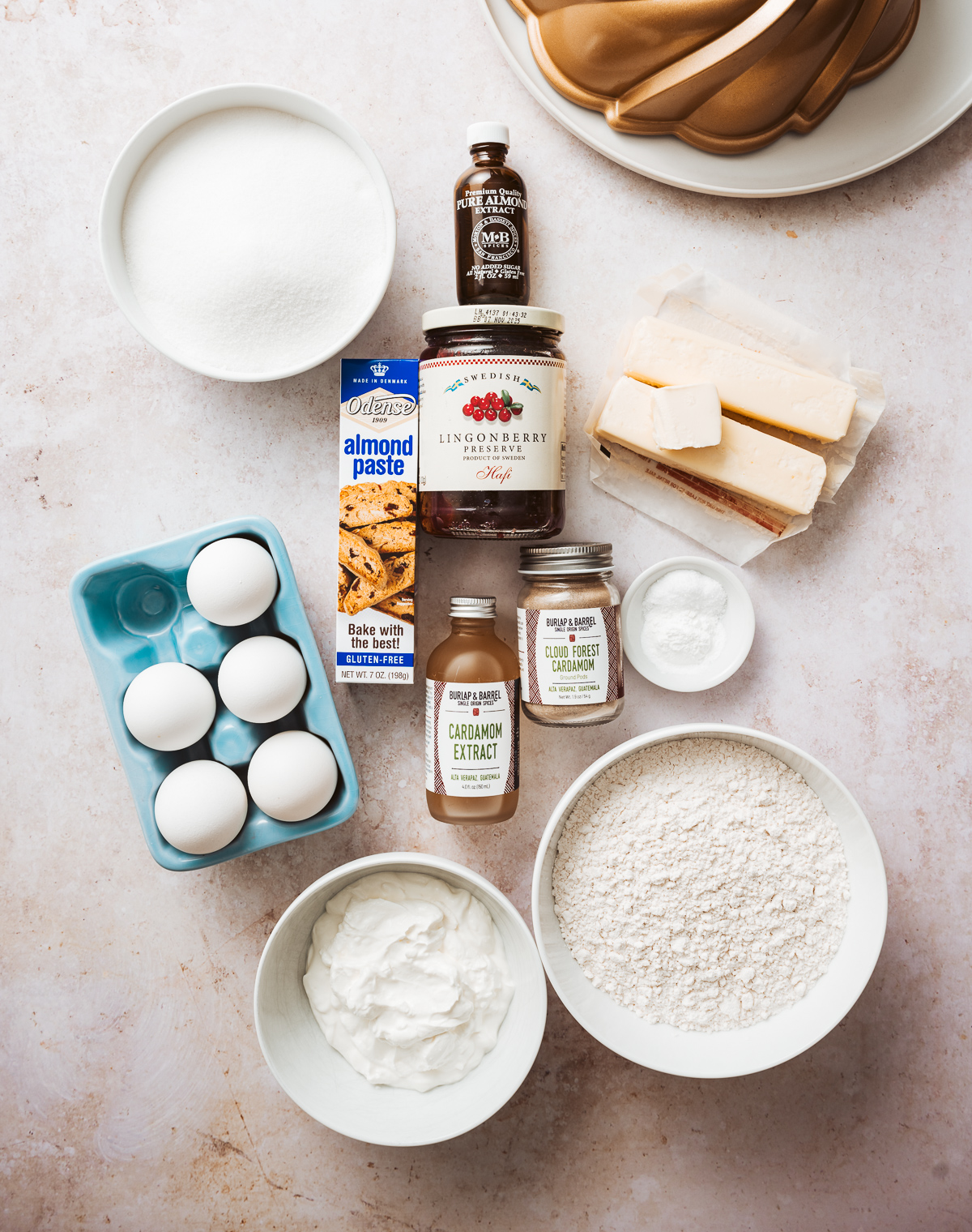 ingredients for cardamom bundt cake eggs almond paste jar of lingonberry jam bowl of flour bowl of sour cream butter jar of cardamom extract jar of ground cardamom