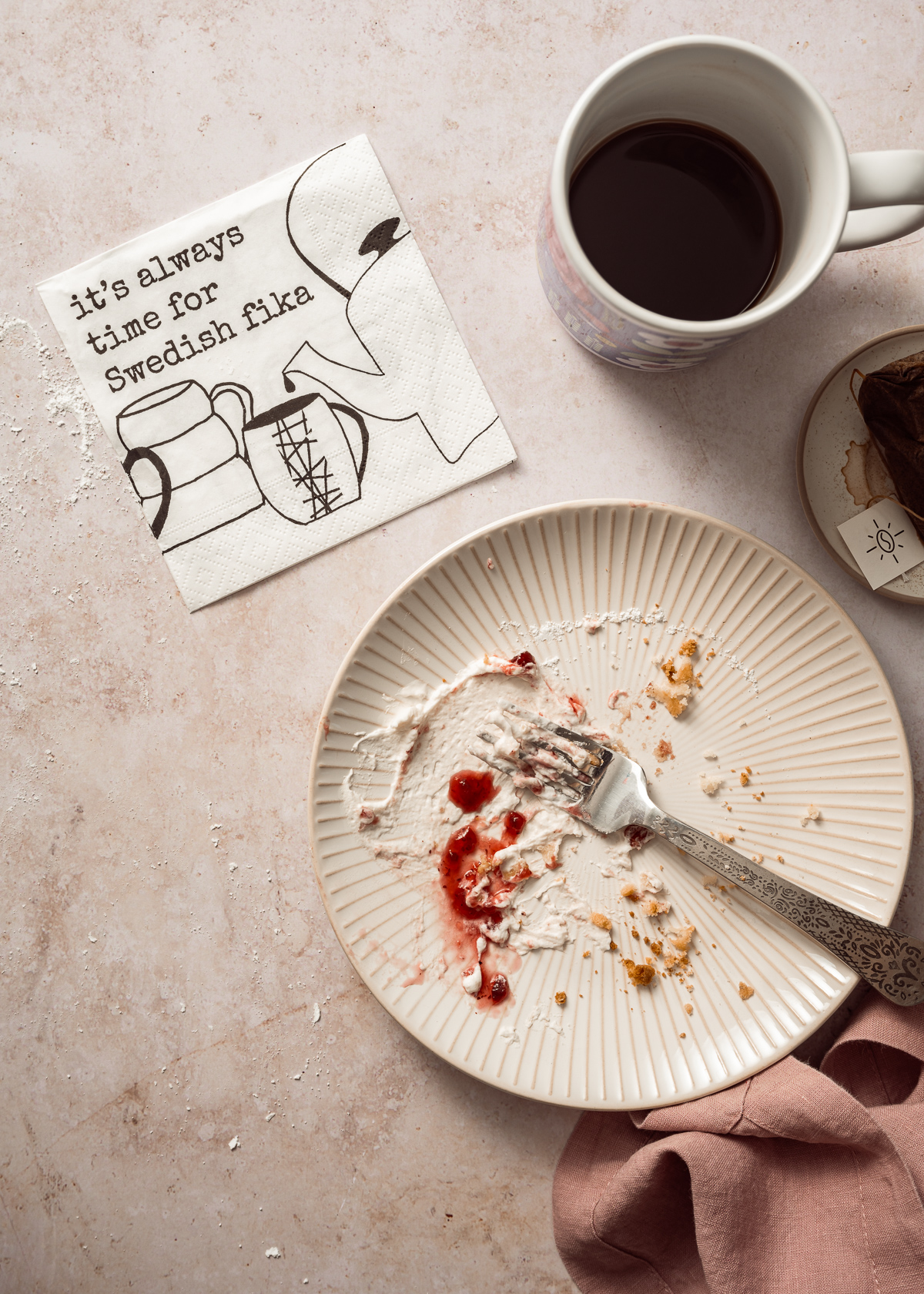 coffee cup, plate with a few cake crumbs a some whipped cream and a smear of lingonberry jam fork pink napkin printed napkin