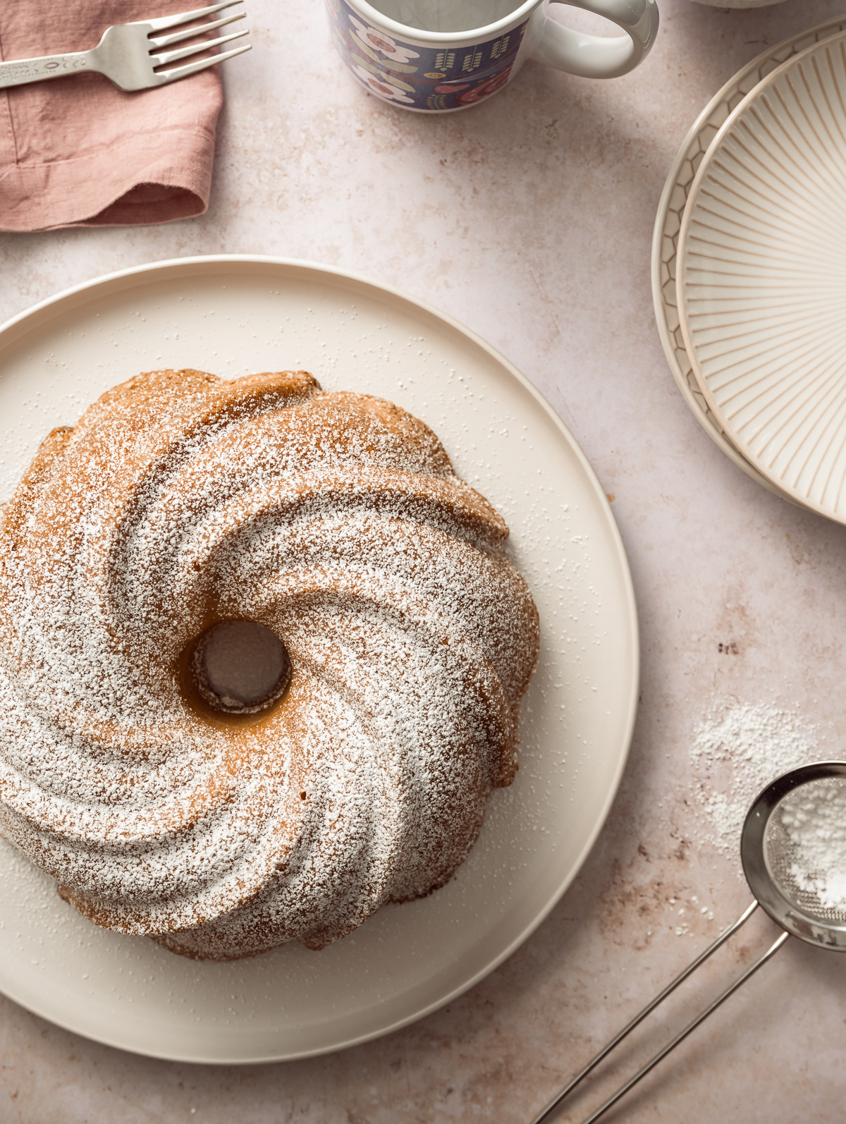 bundt cake on platter dusted with powdered sugar plates coffee cup fork pink napkin strainer with powdered sugar