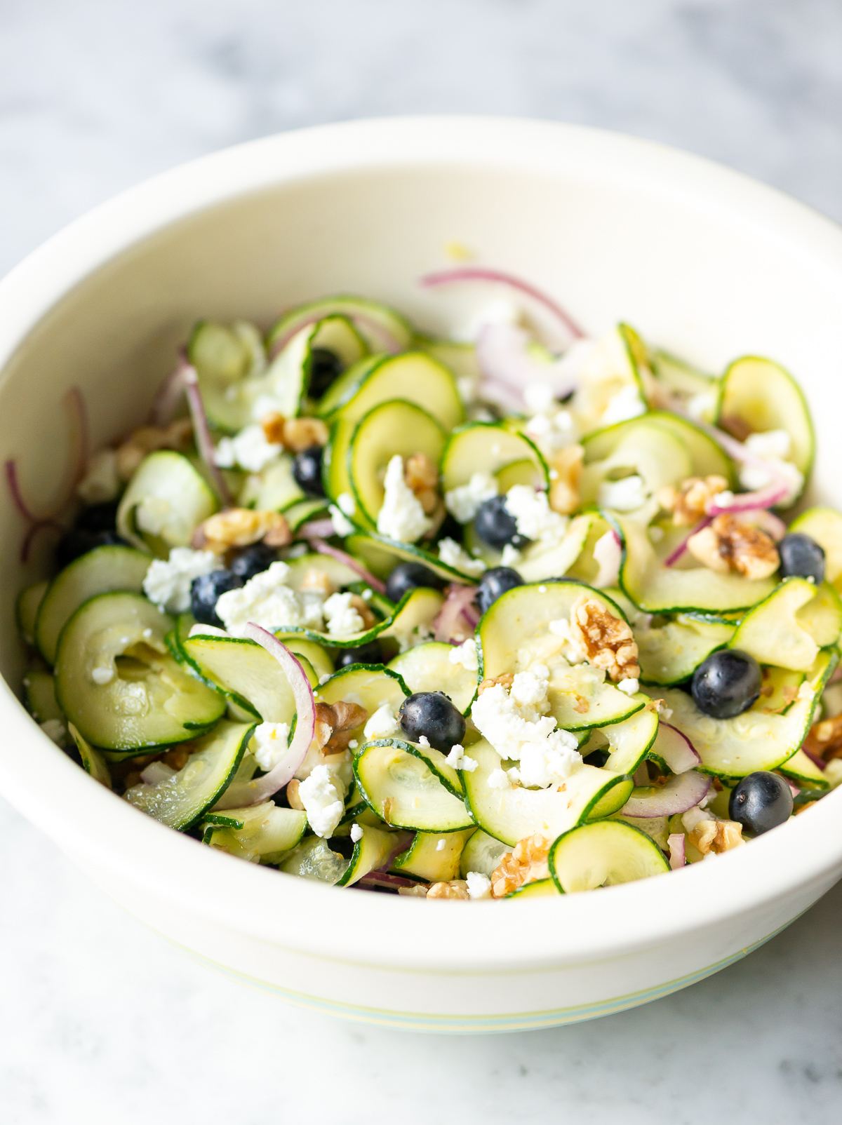 bowl full of zucchini ribbon salad with red onion slices blueberries walnut pieces and goat cheese crumbles