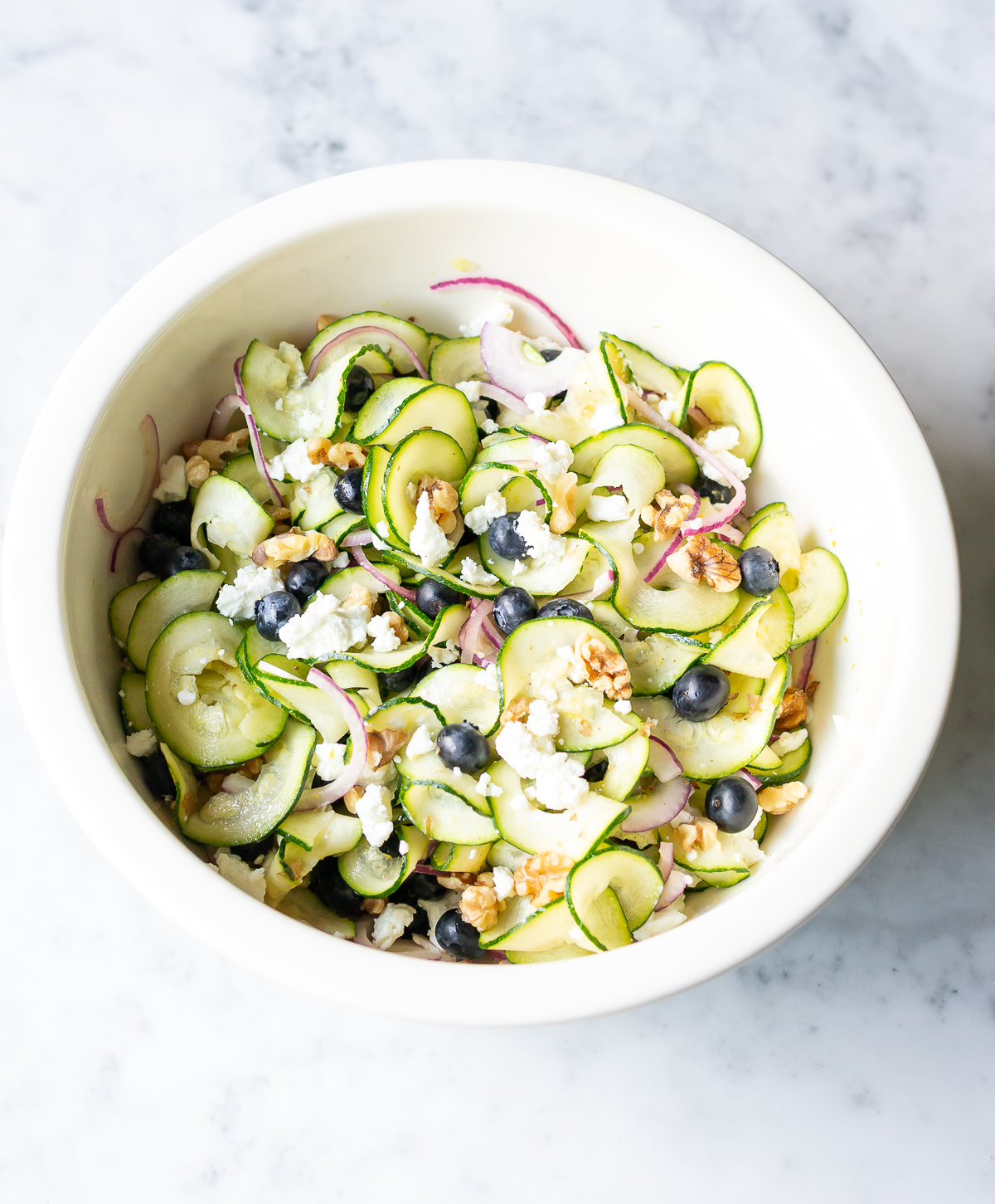bowl full of zucchini ribbons salad with red onion slices blueberries walnut pieces and goat cheese crumbles