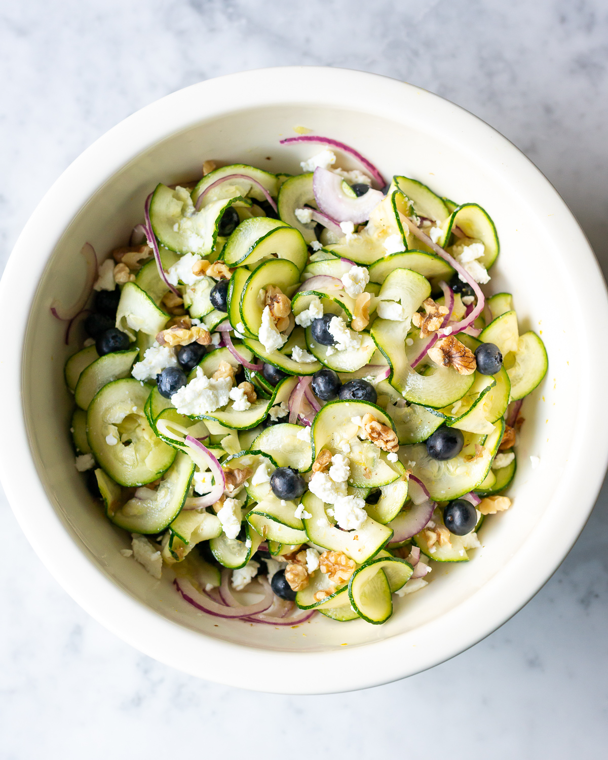 bowl full of zucchini ribbons salad with red onion slices blueberries walnut pieces and goat cheese crumbles