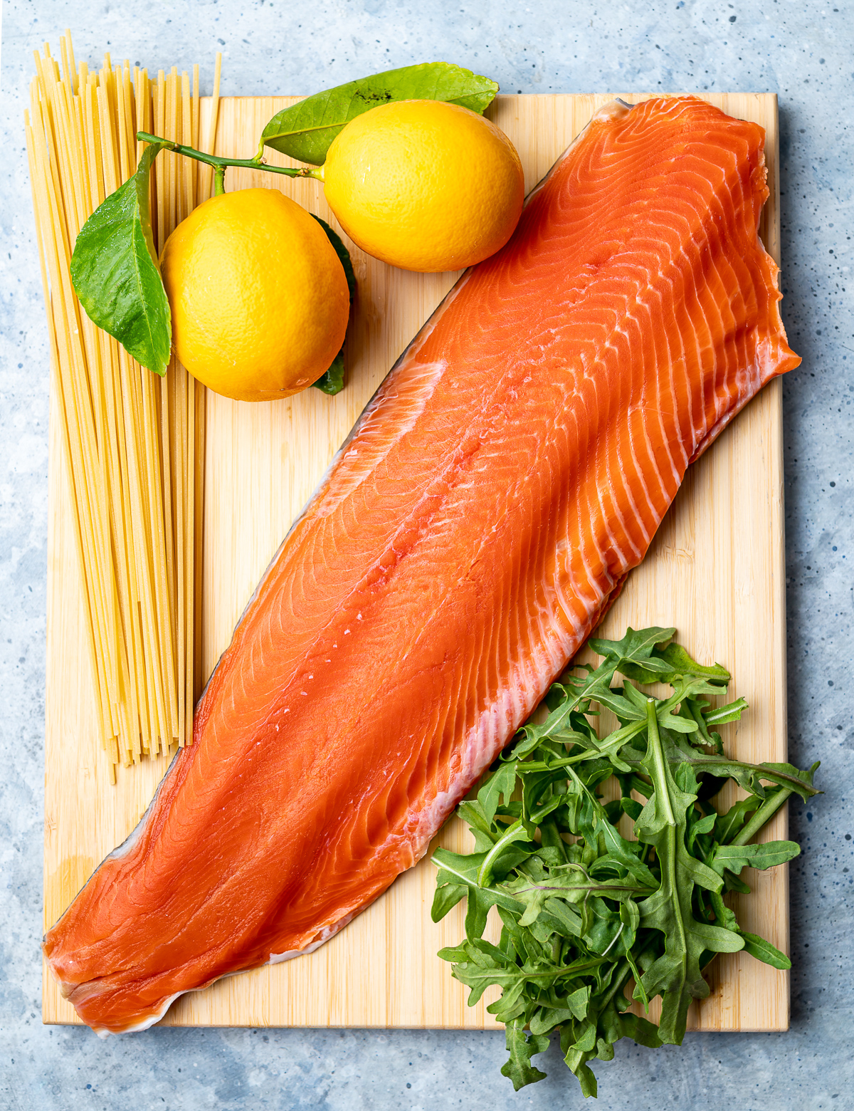 salmon fillet on wood board with fresh arugula a bunch of dried spaghetti noodles and two whole meyer lemons 